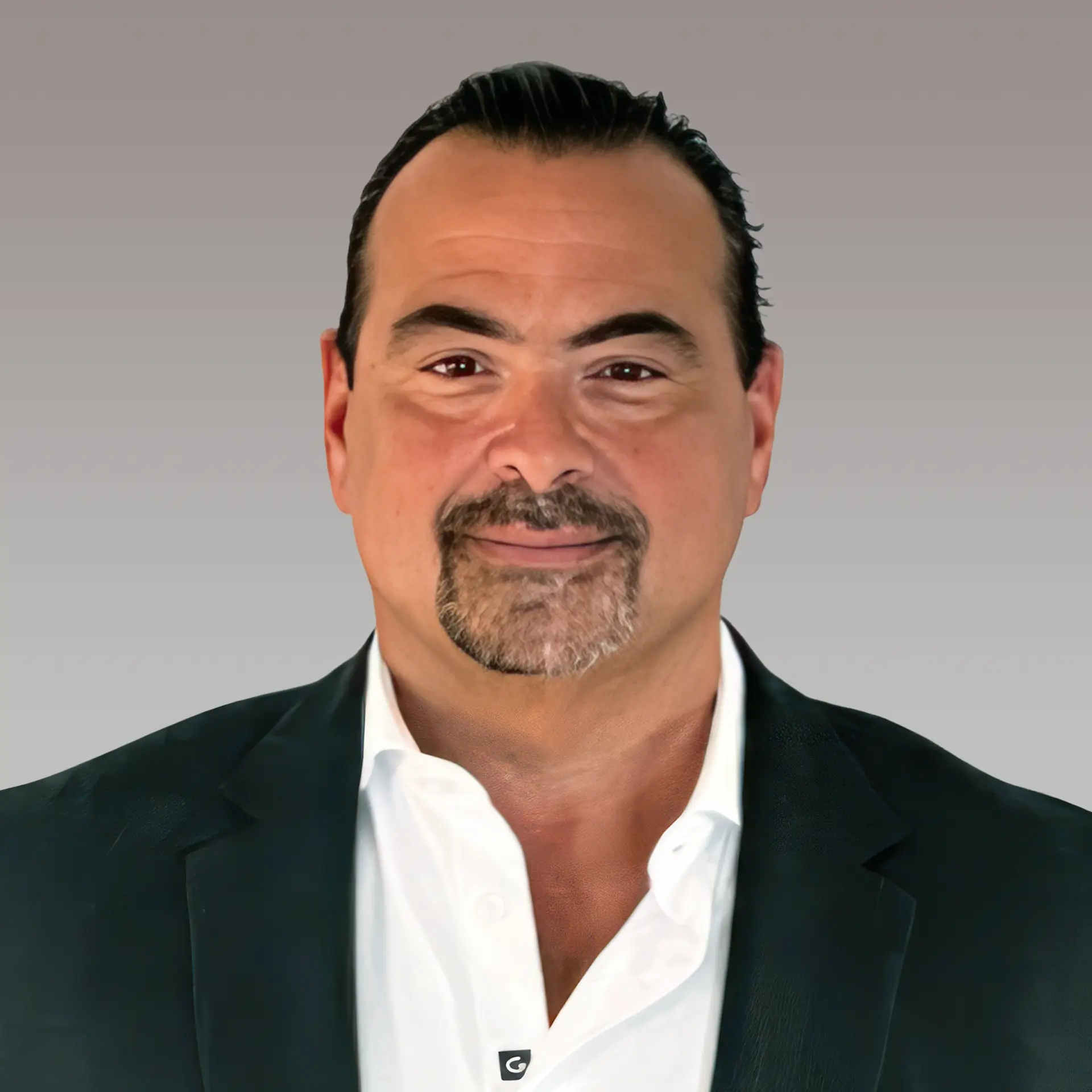 A man with short dark hair and a goatee is wearing a black blazer over a white shirt. He is smiling slightly against a plain gray background.