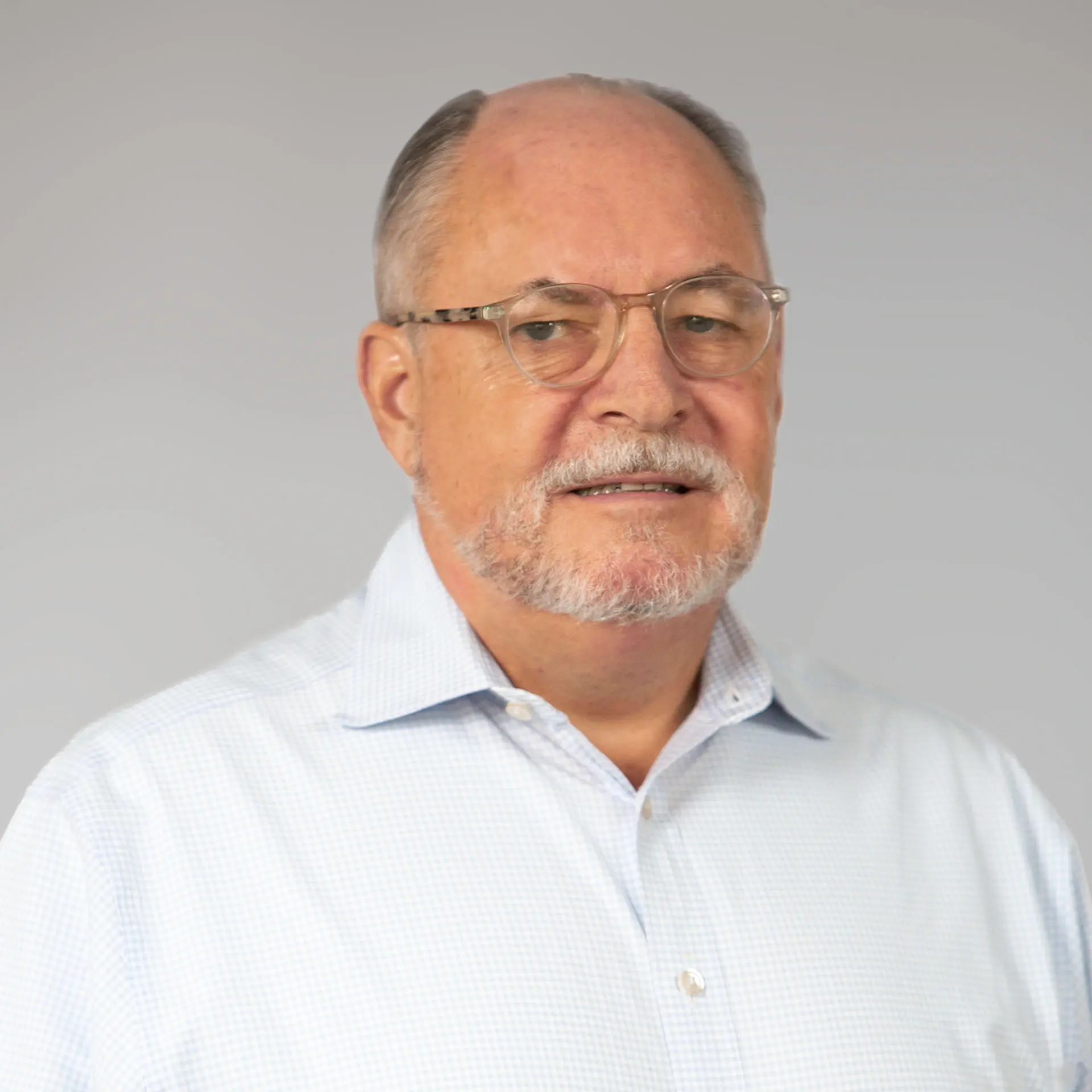 A man with a gray beard and glasses wearing a light blue collared shirt poses for a portrait against a plain background.