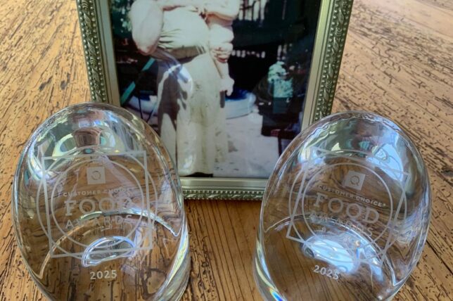 A framed photo of a woman holding a child stands behind two clear glass awards labeled Food and dated 2023, all placed on a wooden table.